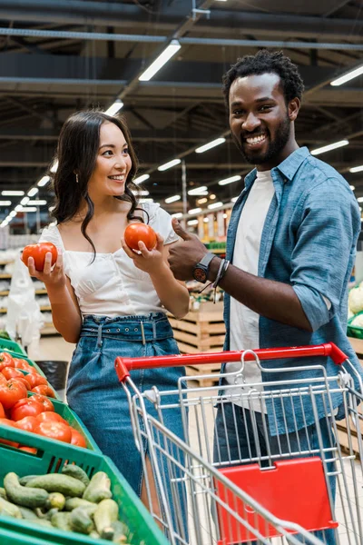 Feliz asiático mujer holding fresco tomates cerca africano americano hombre mostrando pulgar arriba en supermercado - foto de stock