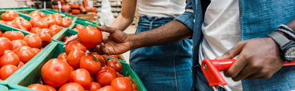 Plano panorámico de hombre afroamericano sosteniendo tomate fresco cerca de chica en el supermercado - foto de stock
