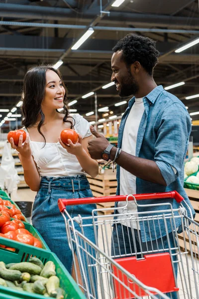 Selective focus of happy asian woman holding fresh tomatoes near cheerful african american man showing thumb up in supermarket — Stock Photo