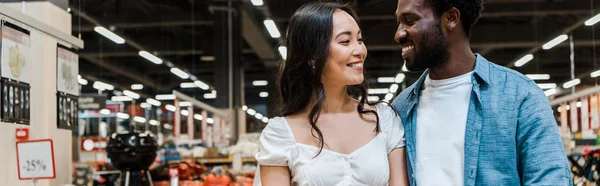 Tiro panorámico de hombre afroamericano feliz mirando atractiva mujer asiática - foto de stock