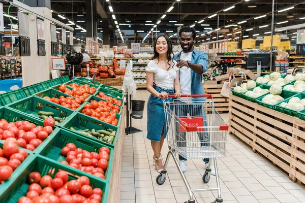 Foyer sélectif de heureuse femme asiatique près de l'homme afro-américain pointant du doigt les tomates dans le supermarché — Photo de stock