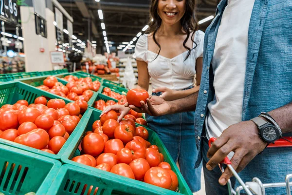 Vista recortada de la mujer alegre limpio afroamericano hombre sosteniendo tomate - foto de stock