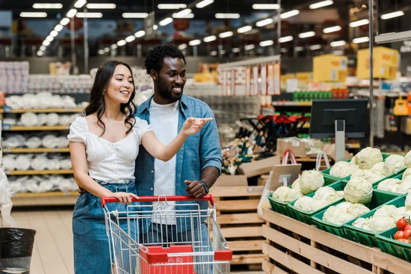 Cheerful african american man near happy asian woman pointing with finger in supermarket — Stock Photo