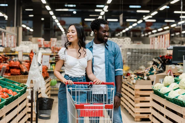 Fröhlicher afrikanisch-amerikanischer Mann und glückliche asiatische Frau in der Nähe von Einkaufswagen im Supermarkt — Stockfoto