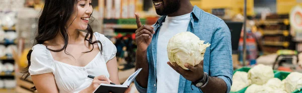 Panoramic shot of happy african american man gesturing and holding cabbage near cheerful asian woman with notebook — Stock Photo