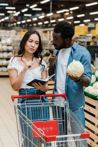 Handsome african american man holding cabbage and pointing with finger at notebook near asian woman — Stock Photo