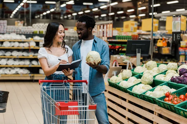 Cheerful african american man holding cabbage and looking at asian woman with notebook and pen — Stock Photo
