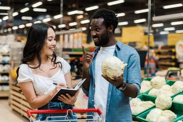 Happy african american man gesturing and holding cabbage near cheerful asian woman with notebook — Stock Photo