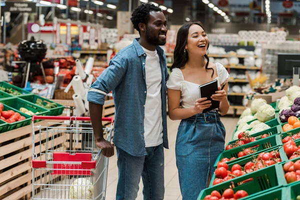 Веселий афроамериканський чоловік біля привабливої азіатської жінки тримає блокнот у супермаркеті — стокове фото
