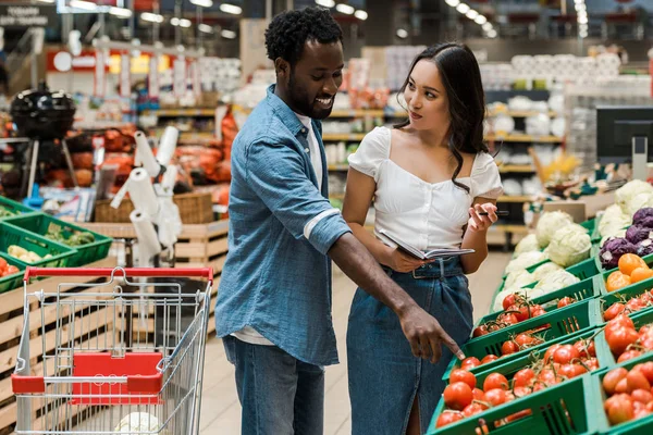Alegre africano americano mn apuntando con dedo en orgánico tomates cerca feliz asiático mujer - foto de stock