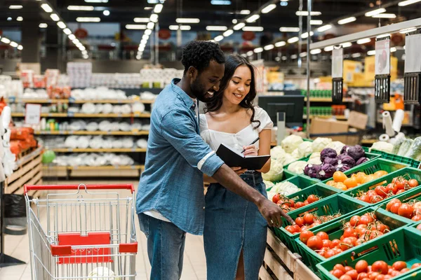 Alegre africano americano mn apuntando con dedo en maduro tomates cerca feliz asiático mujer - foto de stock