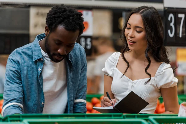 Asiático mulher com notebook de pé perto africano americano homem no supermercado — Fotografia de Stock