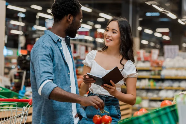 Afrikanisch-amerikanischer Mann hält frische Tomaten in der Hand und schaut fröhliche asiatische Frau mit Notizbuch an — Stockfoto