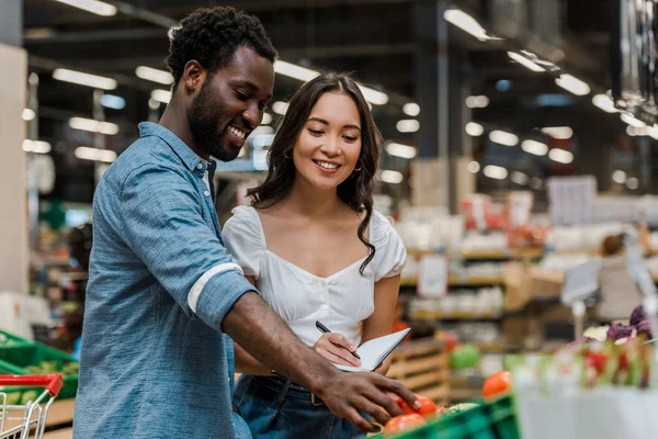 Enfoque selectivo de hombre afroamericano feliz tocando tomates rojos cerca de la mujer asiática en la tienda - foto de stock