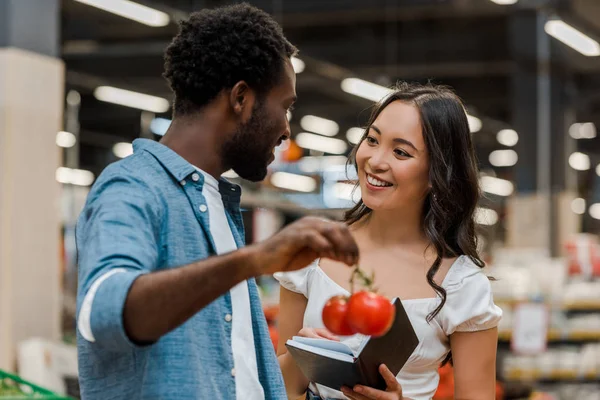 African american man holding fresh tomatoes and looking at happy asian girl with notebook in store — Stock Photo