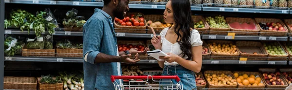 Colpo panoramico di felice uomo africano americano mostrando gesto scrollata di spalle vicino donna asiatica con notebook che punta con il dito — Foto stock
