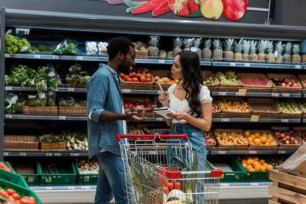 Feliz africano americano hombre mostrando encogiéndose de hombros gesto cerca asiático mujer con notebook apuntando con dedo - foto de stock