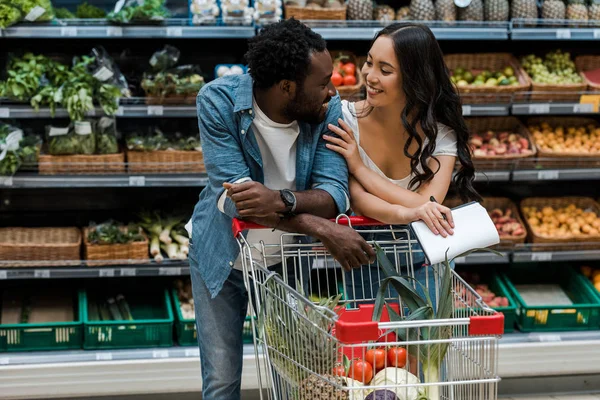 Feliz homem americano africano e mulher asiática em pé perto de carrinho de compras no supermercado — Fotografia de Stock