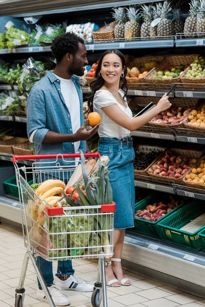 Feliz homem americano africano olhando para mulher asiática em pé no supermercado perto de frutas — Fotografia de Stock