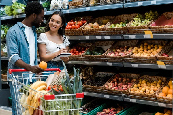 Glücklicher afrikanisch-amerikanischer Mann schaut asiatische Frau an, die mit Notizbuch und Stift im Supermarkt steht — Stockfoto