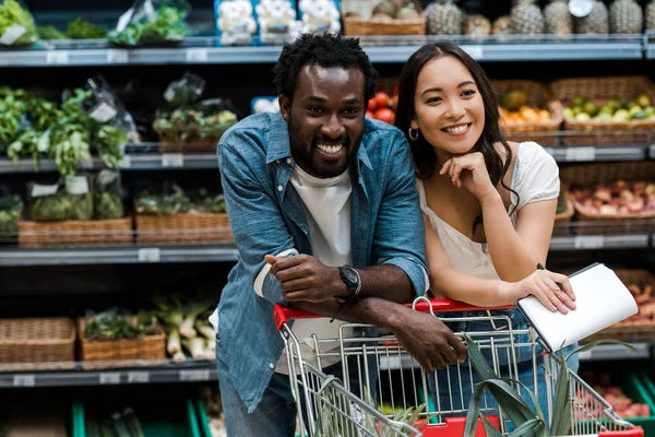 Alegre pareja interracial sonriendo en supermercado cerca de carrito de compras - foto de stock
