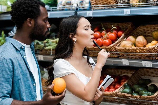 Pensive asian woman with pen standing near fruits and african american man with orange — Stock Photo
