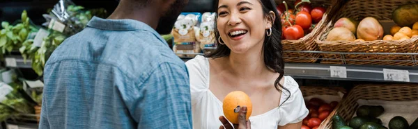 Панорамный снимок счастливой азиатской девушки рядом с африканским американцем с апельсином — стоковое фото