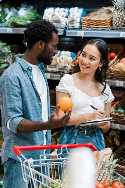 Enfoque selectivo de feliz chica asiática cerca de hombre afroamericano con naranja - foto de stock