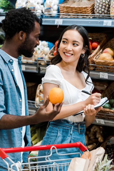 Selective focus of happy asian girl with notebook and pen near cheerful african american man with orange — Stock Photo