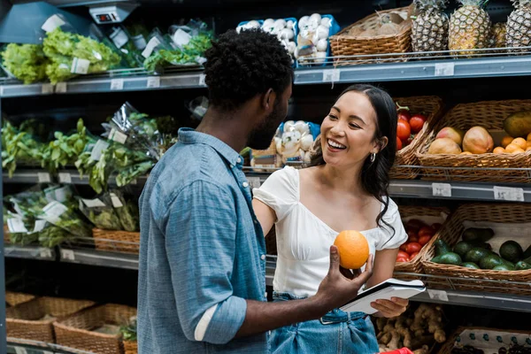 Feliz asiático chica cerca africano americano hombre con naranja en tienda - foto de stock
