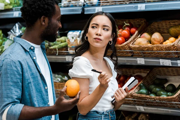 Pensivo asiático menina olhando para feliz africano americano homem com laranja — Fotografia de Stock