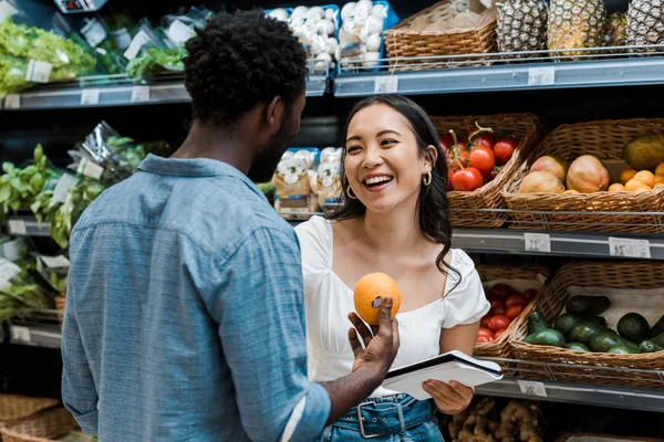 Alegre asiático chica cerca africano americano hombre con naranja en tienda - foto de stock