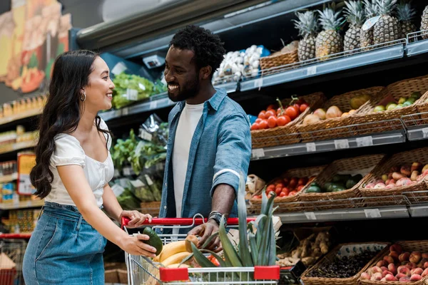 Happy asian girl holding avocado near shopping cart and african american man in store — Stock Photo