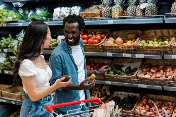 Happy asian girl holding avocado near shopping cart and african american man with kiwi fruit in supermarket — Stock Photo