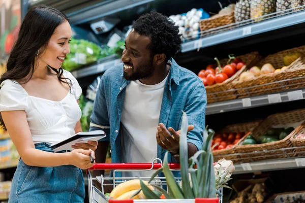 Selective focus of happy african american man gesturing and looking at asian girl holding notebook — Stock Photo