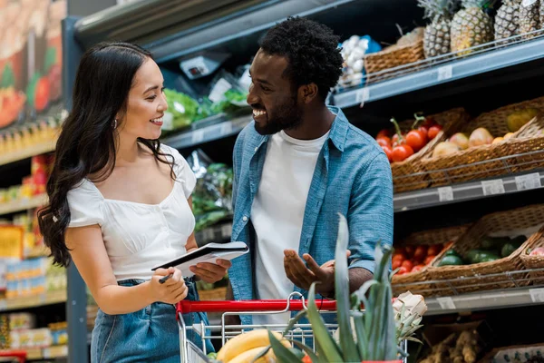 Selektiver Fokus eines glücklichen afrikanisch-amerikanischen Mannes, der gestikuliert und ein asiatisches Mädchen anschaut, das ein Notizbuch im Laden hält — Stockfoto