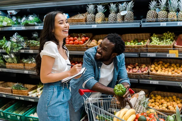 Glücklich afrikanisch-amerikanischer Mann schaut asiatische Frau mit Notizbuch in der Nähe von Einkaufswagen mit Lebensmitteln an — Stockfoto