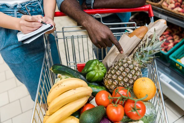 Vista recortada de hombre afroamericano señalando con el dedo en el carrito de la compra con comestibles cerca de la mujer con cuaderno - foto de stock