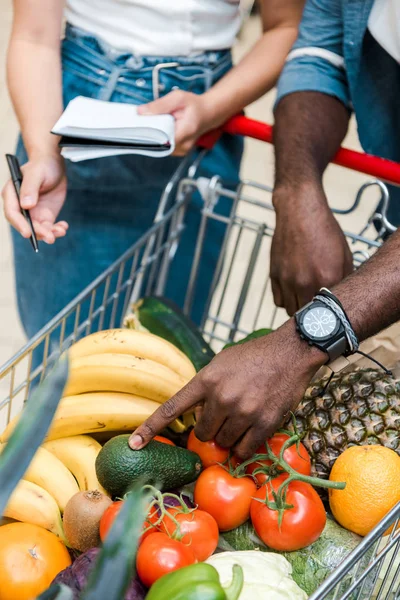 Messa a fuoco selettiva dell'uomo afroamericano che punta con il dito al carrello della spesa con generi alimentari vicino alla donna con notebook — Foto stock