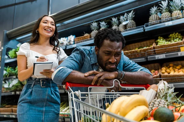 Enfoque selectivo de triste afroamericano hombre mirando el carrito de compras cerca de feliz mujer asiática con portátil - foto de stock