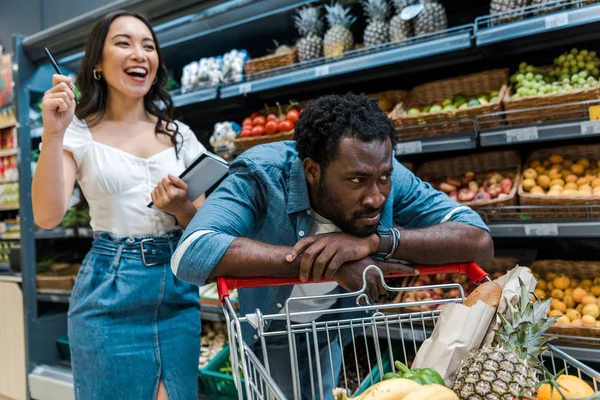 Selective focus of sad african american man near shopping cart near happy asian woman with notebook — Stock Photo