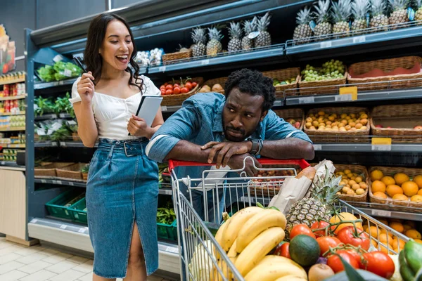 Selective focus of sad african american man near shopping cart near happy asian woman with notebook — Stock Photo