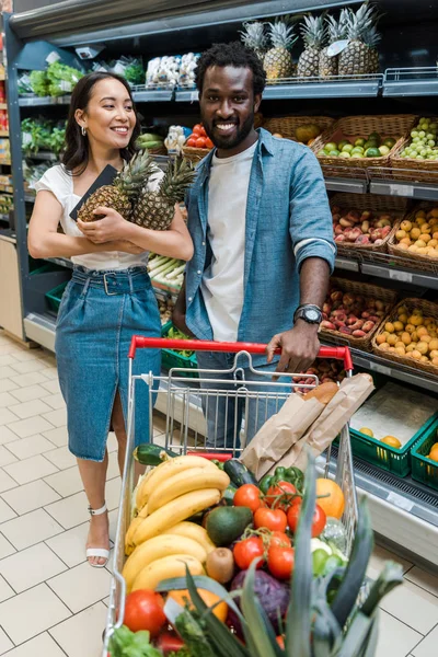 Selective focus of happy asian woman holding pineapples near african american man in supermarket — Stock Photo