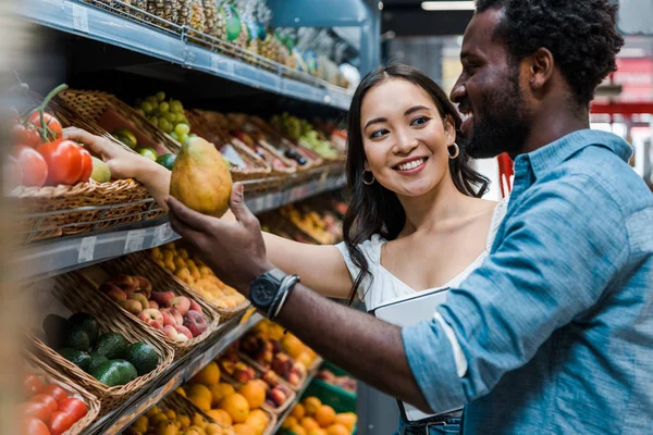 Feliz mulher asiática olhando para o homem americano africano perto de frutas na loja — Fotografia de Stock