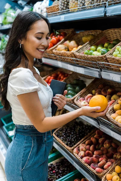 Selective focus of cheerful asian woman looking at orange near fruits in store — Stock Photo