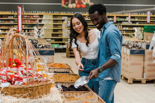 Glücklich afrikanisch-amerikanischer Mann hält Metallschaufel, während er Rosinen in der Nähe einer asiatischen Frau im Geschäft nimmt — Stockfoto