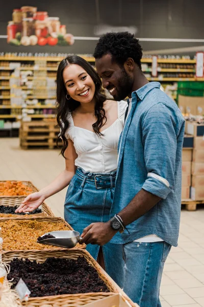 Heureux afro-américain homme tenant cuillère en métal avec raisins secs près asiatique femme geste en magasin — Photo de stock