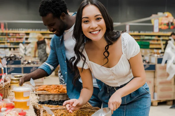 Happy asian woman looking at camera near cheerful african american man looking at honey jars in supermarket — Stock Photo