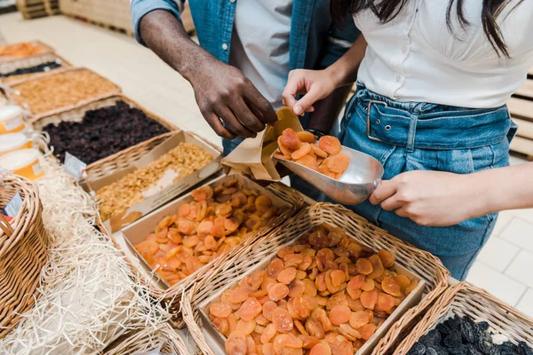 Vue recadrée d'une femme tenant une cuillère en métal avec des abricots secs près d'un homme afro-américain avec un sac en papier — Photo de stock