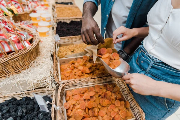 Vista ritagliata di donna in possesso di paletta di metallo con albicocche secche vicino all'uomo africano americano con sacchetto di carta nel supermercato — Foto stock